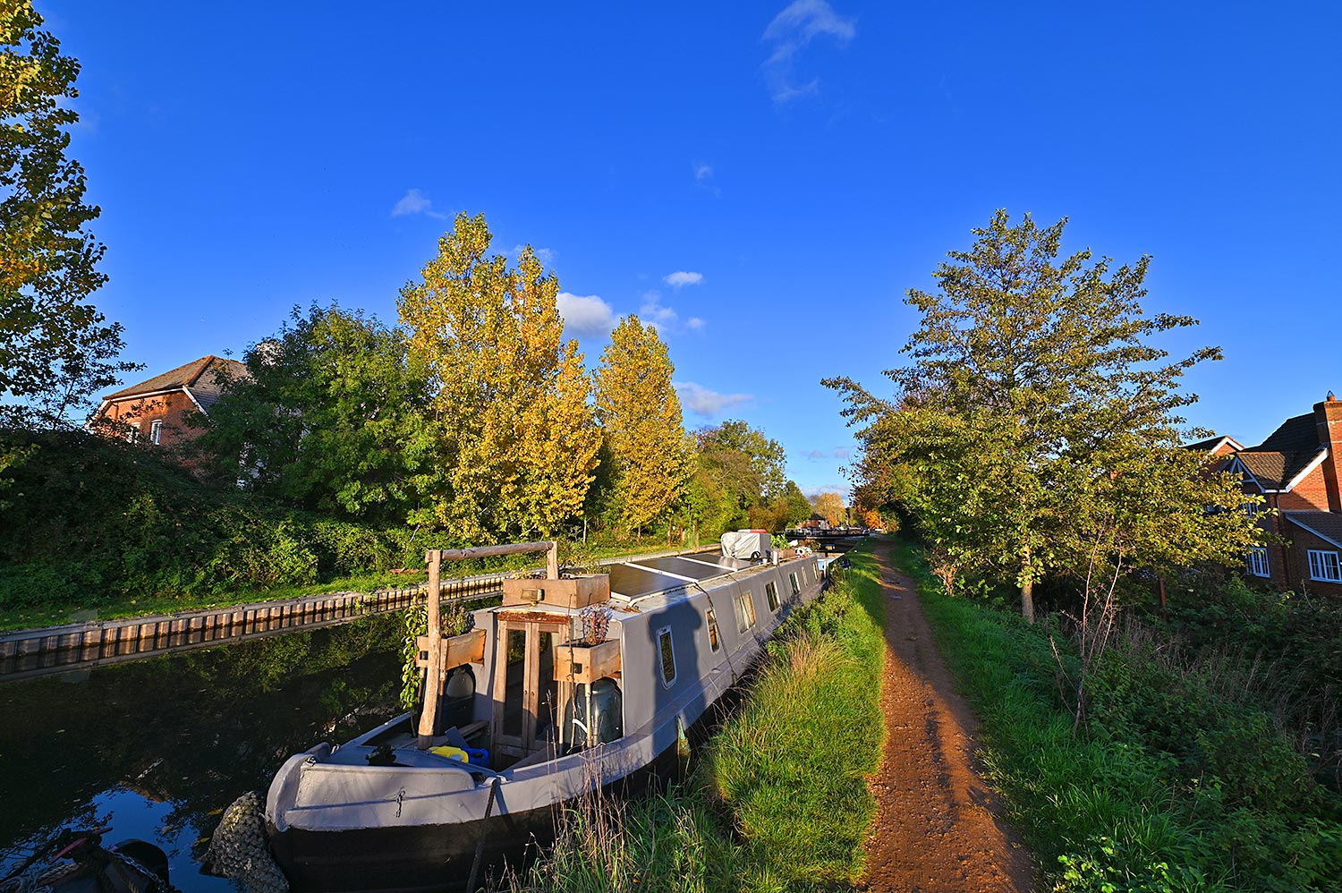 Picture of a canal boat moored along a canal in the autumn sunshine, trees in their autumn colour in the background